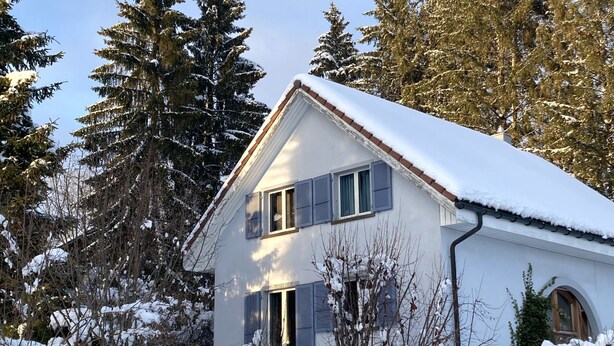 Ein weißes Haus mit blauen Fensterläden, umgeben von schneebedeckten Bäumen und wolkenlosem Himmel.