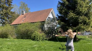 Eine Person spannt einen Bogen in einer grünen Landschaft, im Hintergrund ist ein Haus zu sehen.
