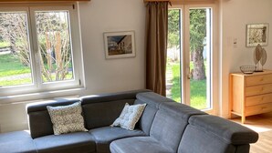 Ein modernes Wohnzimmer mit einem grauen Ecksofa, einer Holzkommode und einem großen Fenster mit Blick auf Grünflächen.