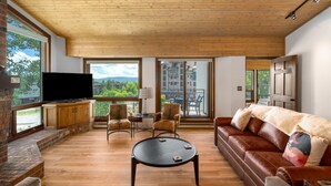 Uma sala de estar espaçosa com teto de madeira, uma TV grande, lareira, sofá de couro e janelas amplas com vista para o exterior.