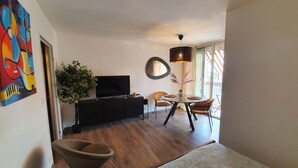 A modern living room with a flat-screen TV on a black TV stand, a dining area with a round table and two chairs, and a large window with a view of the outdoors.
