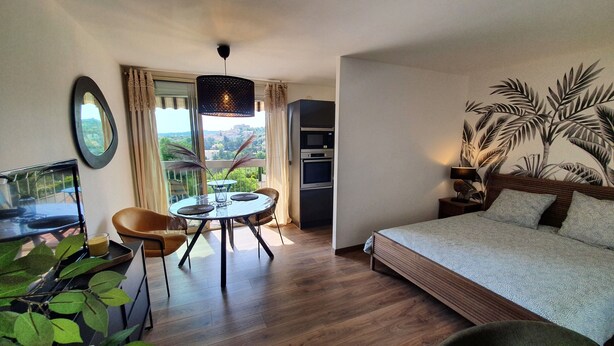 A modern hotel room with a bed, a dining table with chairs, a TV, and a balcony with a view.