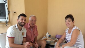 Tre personer sidder på fletstole i et rum med flisegulv og et vindue med metalramme.