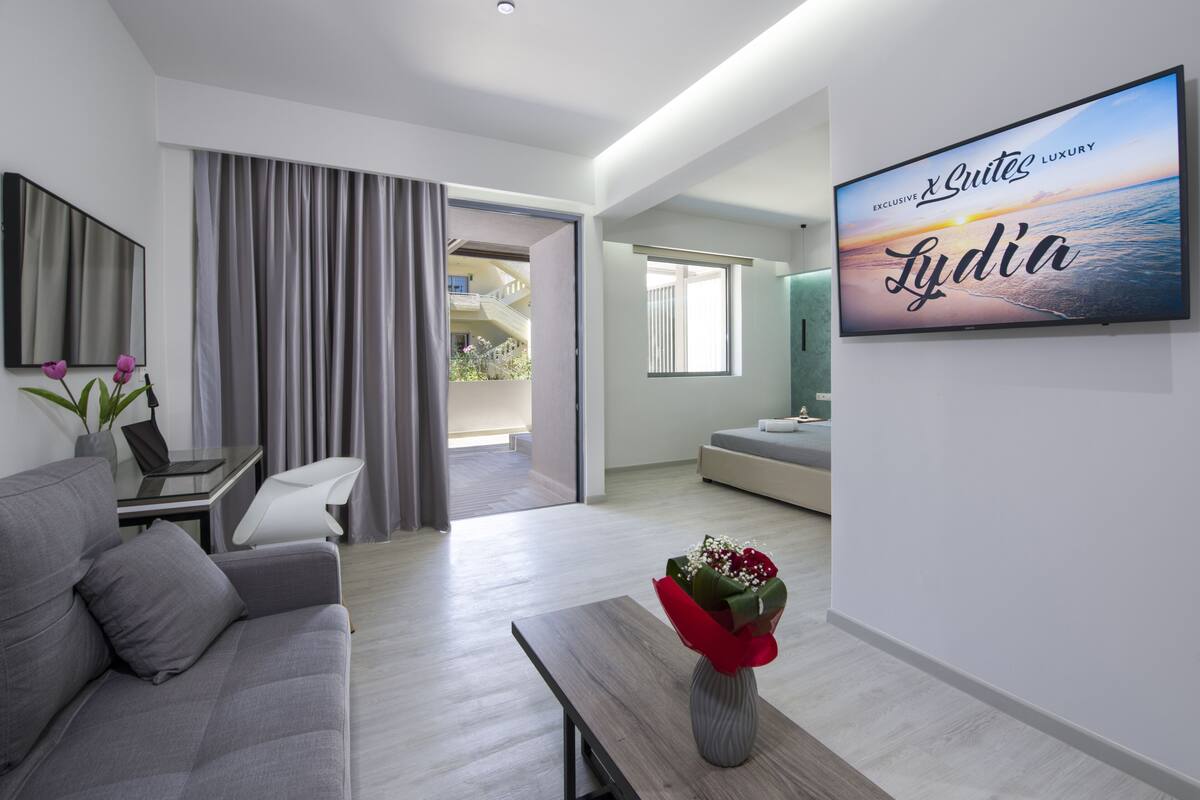 Un moderno cuarto de hotel con un sofá gris, una mesa de centro de madera con un jarrón rojo, un televisor de pantalla plana en la pared y vista al mar por la ventana.