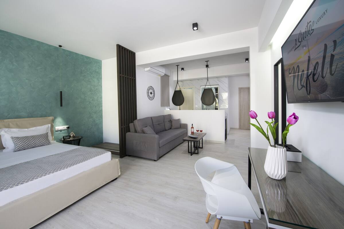 Un dormitorio moderno con cama, un sofá gris, una mesita con una botella y vasos, y una silla blanca.