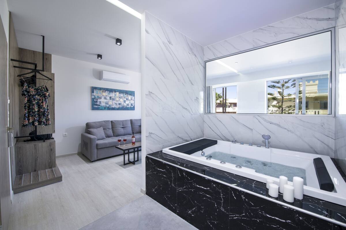 Un baño moderno con un espejo grande, una bañera de mármol negro y un sofá gris en la zona de estar contigua.