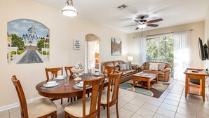 A spacious living room with a large window, a ceiling fan, and a dining area with a round table and chairs.