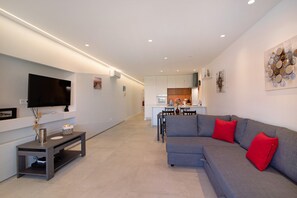 A modern living room with a grey sofa, red cushions, a flat-screen TV, and a dining area in the background.