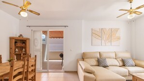 A living room with a sofa, wooden dining table, chairs, ceiling fans, and a sliding glass door leading to a balcony.