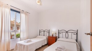 A room with two single beds, a wooden nightstand with a lamp, a window with curtains, and a view of buildings outside.
