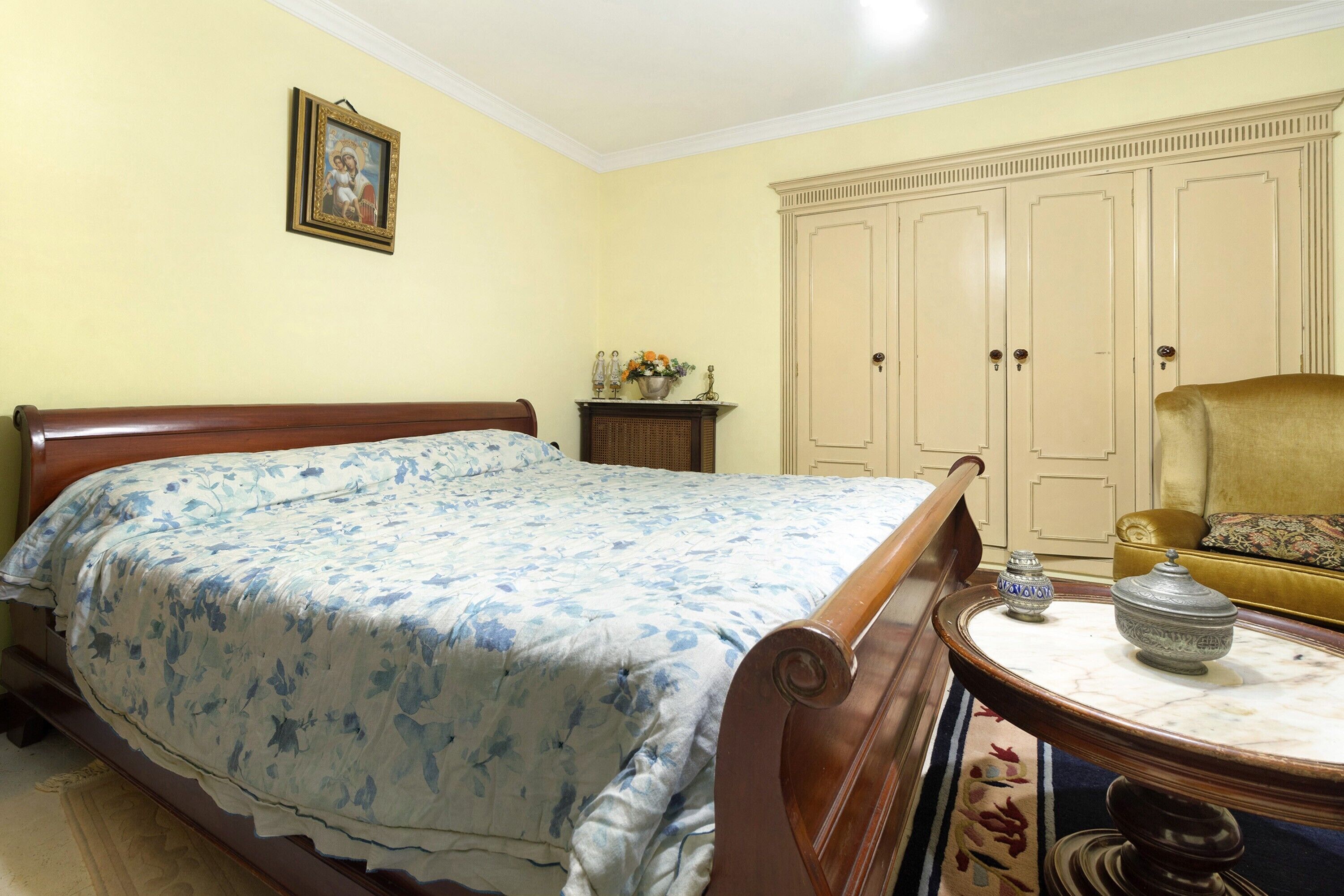 Een slaapkamer met een bed, een dressoir, een stoel en een bijzettafeltje met decoratieve objecten.