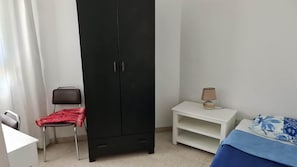 Habitació amb un armari negre, una cadira amb drap vermell, una tauleta de nit blanca amb una làmpada, i un llit amb un sobre quadrat blau i blanc.