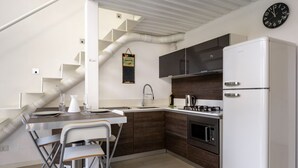 Een moderne keuken met een witte koelkast, fornuis en spoelbak, een eethoek met een tafel en stoelen, en een trap met zichtbare ventilatiekanalen.