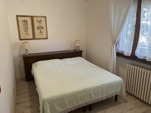 A bedroom with a bed, two bedside lamps, a wooden nightstand, a radiator, and two framed botanical illustrations on the wall. - Apartment (Castiglione della Pescaia)