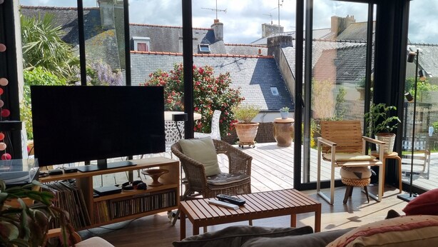 Un salon chaleureux avec un grand téléviseur, des sièges confortables et une vue sur un jardin et les bâtiments voisins.