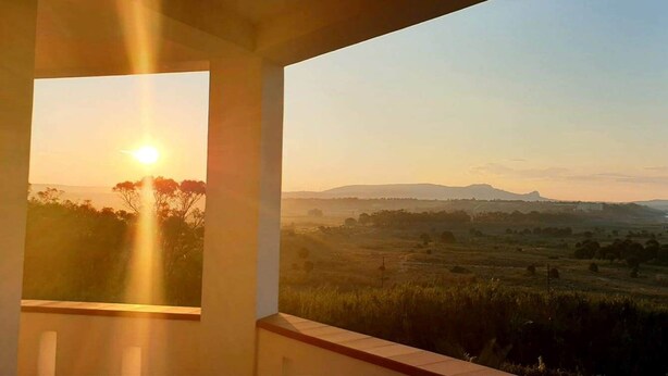 Un balcón con vistas a un atardecer sobre un paisaje con montañas lejanas y una masa de agua.