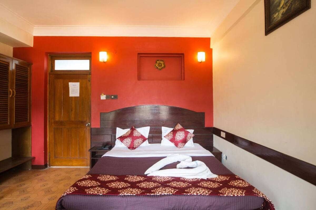 Ein Hotelzimmer mit Holztür, einem Bett mit roten und weißen Kissen, einer Wandleuchte und einem gerahmten Bild an der Wand.