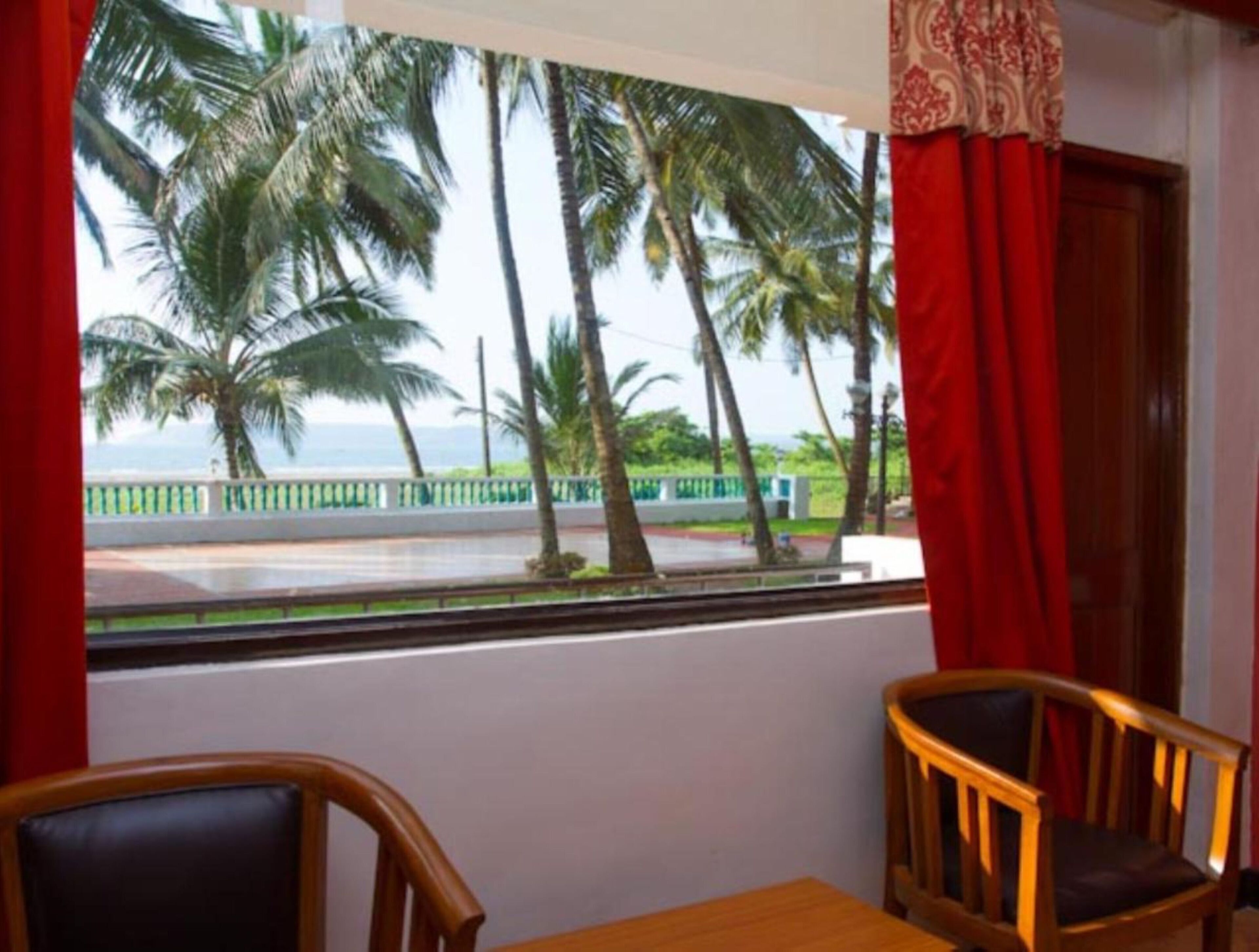 A room with a view of palm trees and a road. There are two wooden chairs and a table.
