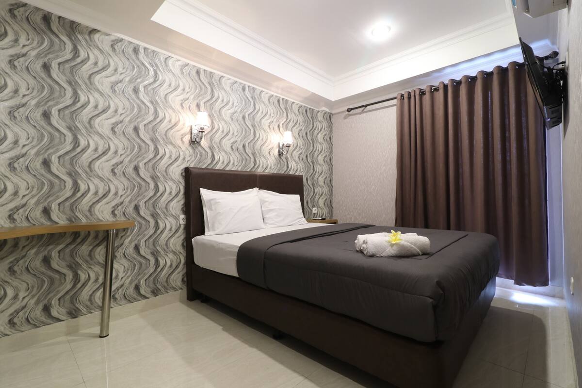 A modern hotel room with a large bed, a wooden side table, and a patterned wallpaper.