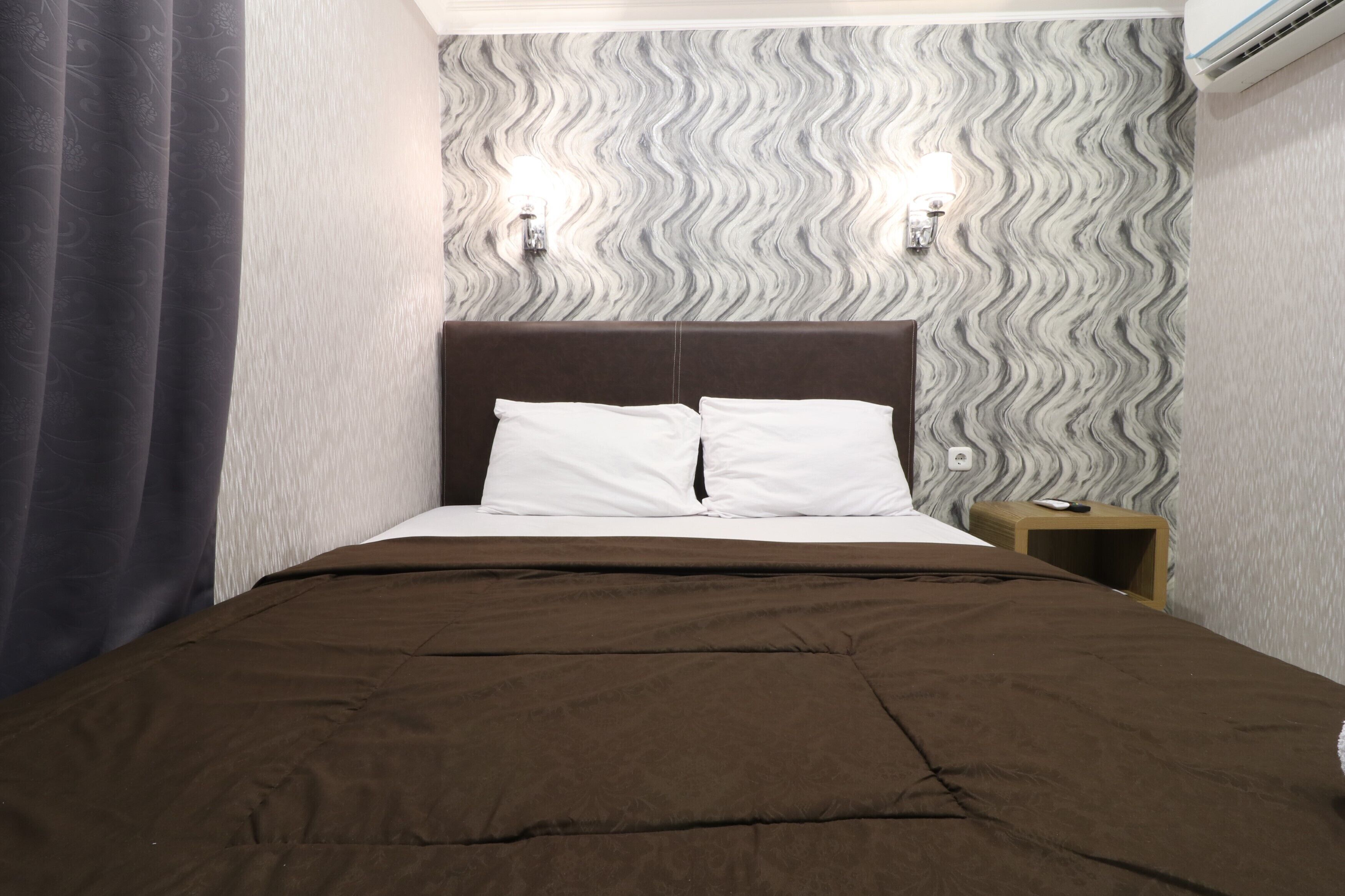 Un dormitorio con cama, cabecera, dos luces fijadas a la pared y una mesita de noche.