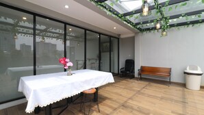 A modern indoor space with a glass wall, a table with a white tablecloth, a bench, and a trash bin. - Flamenco Urban Living (Jakarta)