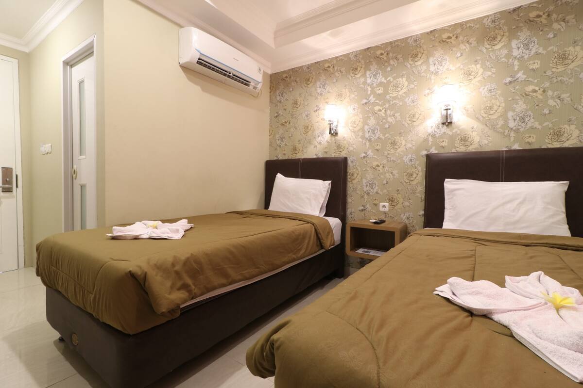 A hotel room with two beds, beige walls, floral wallpaper, and an air conditioning unit.