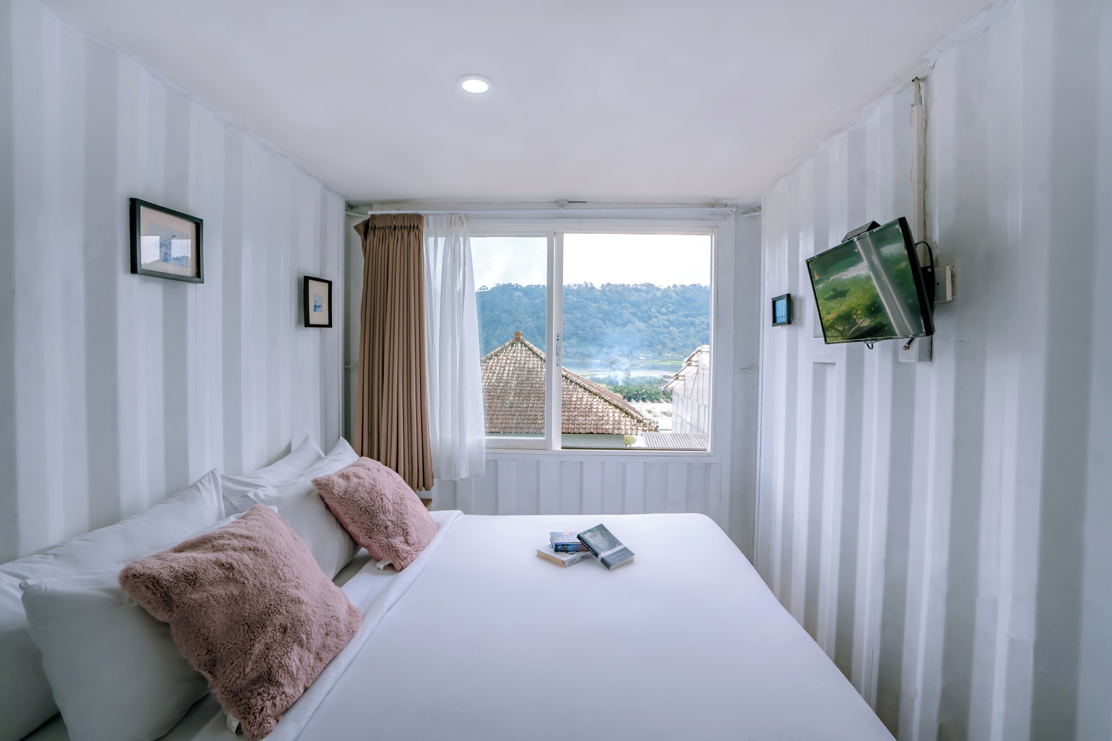 Un dormitorio con cama, almohadas, un televisor montado en la pared y una ventana con vista a edificios y colinas.