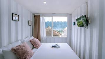 Un dormitorio con cama, almohadas, un televisor montado en la pared y una ventana con vista a edificios y colinas.