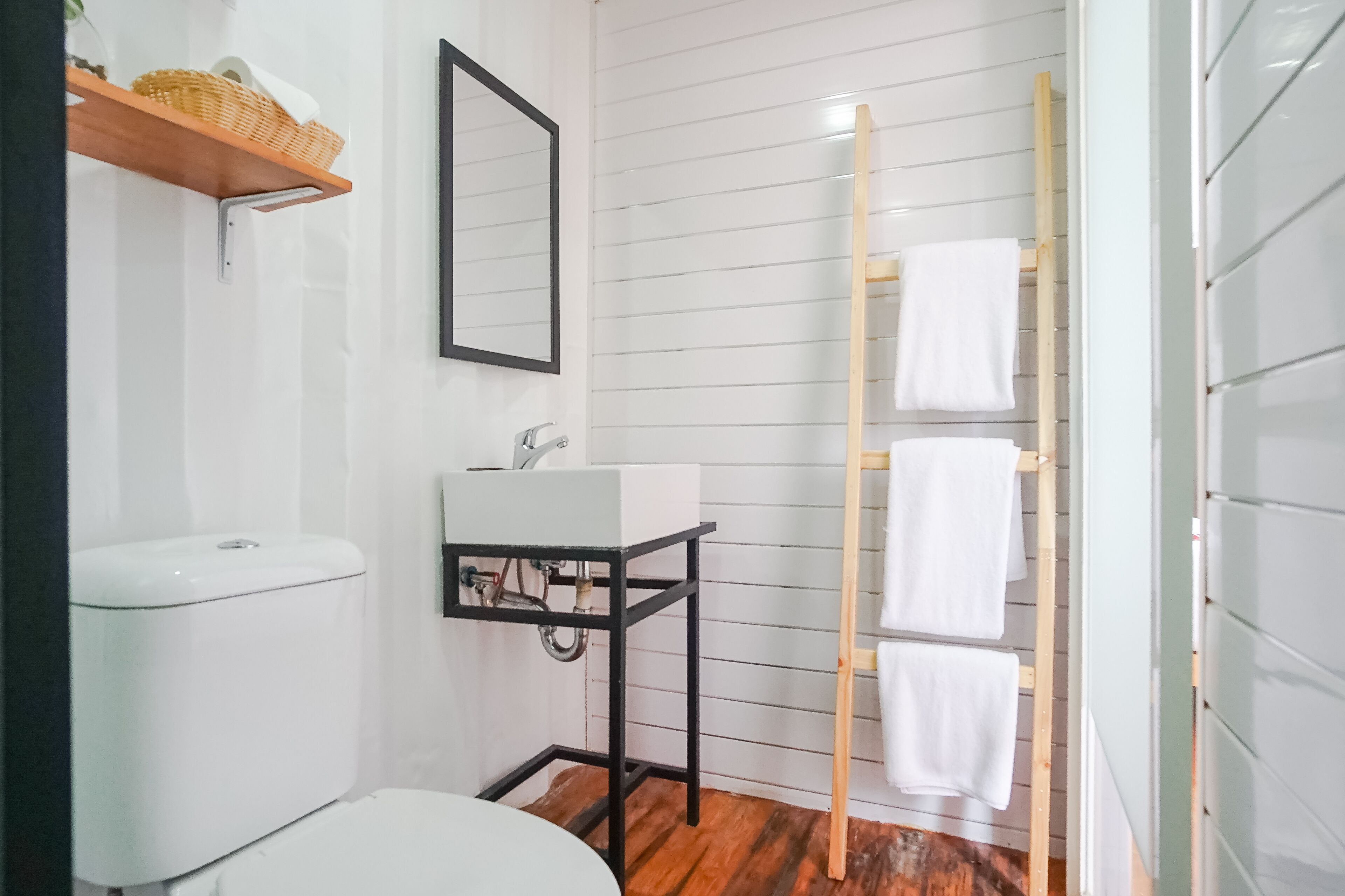 Un baño con un inodoro blanco, un lavamanos blanco sobre un soporte de metal negro, un perchero de madera y una estantería de madera con una cesta.