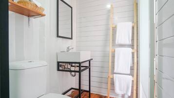 Un baño con un inodoro blanco, un lavamanos blanco sobre un soporte de metal negro, un perchero de madera y una estantería de madera con una cesta.