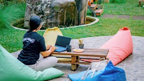 A person sitting on a green bean bag chair using a laptop on a stone patio with a wooden table, surrounded by colorful cushions and a large rock.