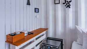 A hotel room with a wooden dresser, a black suitcase, a bed with white bedding, and a wall-mounted shelf with a picture frame and decorative items.