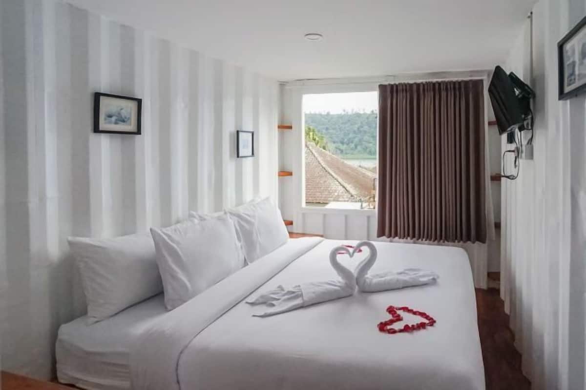 Una habitación de hotel con cama, cortinas blancas, ventanal con vistas y televisor montado.
