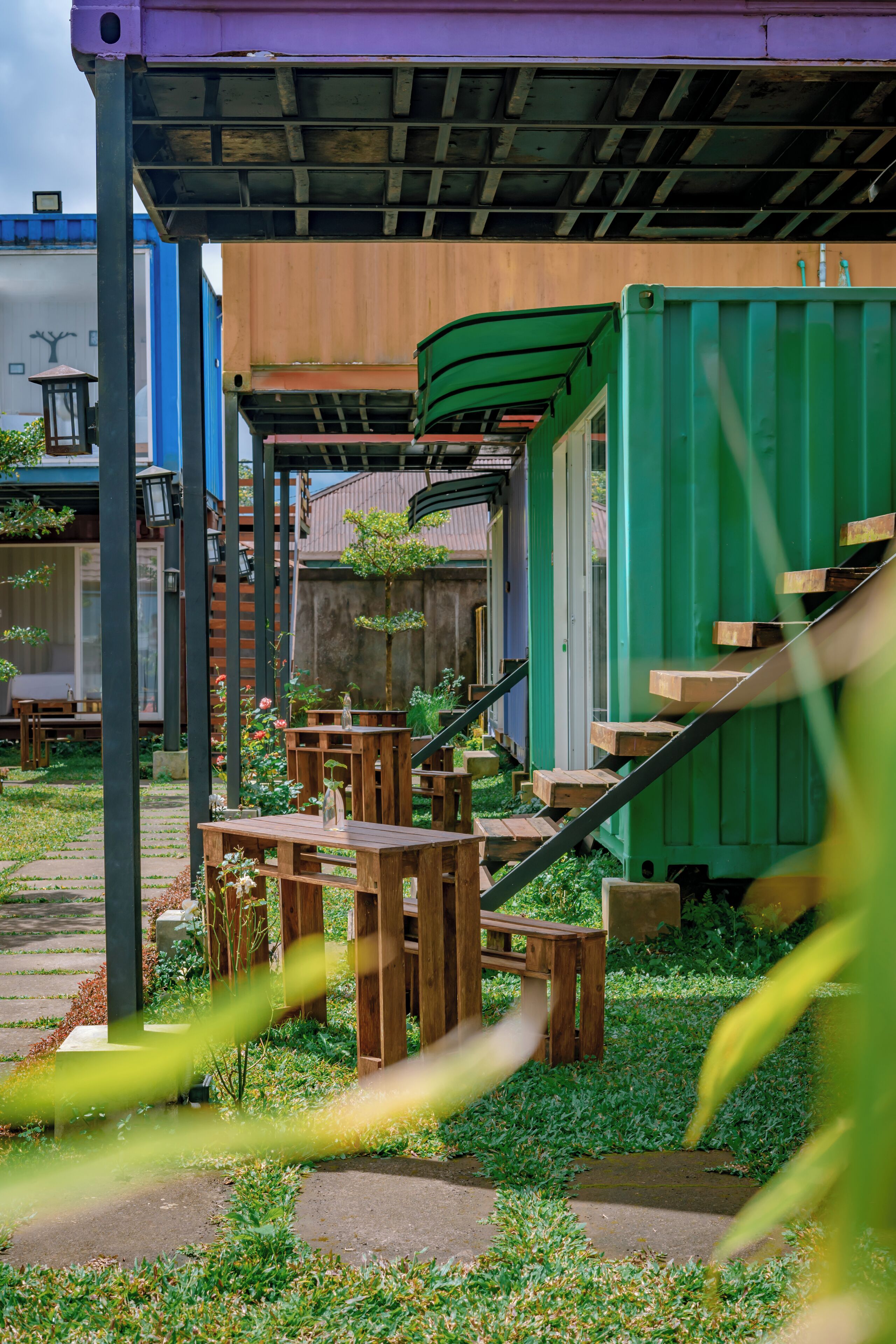A covered outdoor seating area with wooden tables and chairs, a staircase leading to a building, and a green shipping container structure.