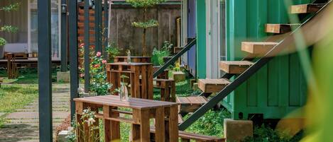 A covered outdoor seating area with wooden tables and chairs, a staircase leading to a building, and a green shipping container structure.