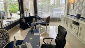 A modern living room with a dark blue sofa, a dining table with chairs, a laptop on a desk, and a large window with a view of parked cars outside. - Roemah Moesi Medan (Medan)