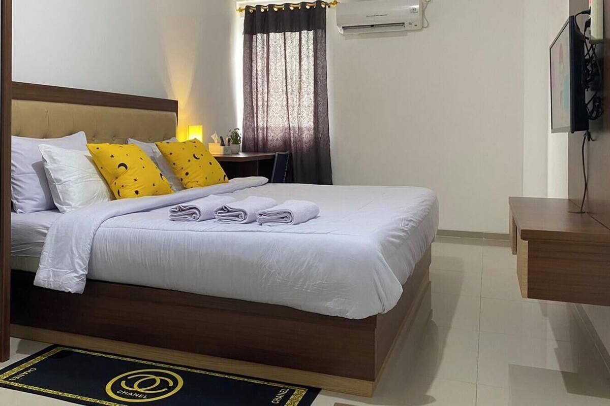 A hotel room with a bed, a desk, a chair, a TV, and an air conditioning unit.