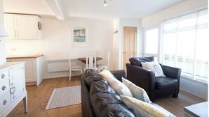Un salon moderne avec un canapé en cuir, une table à manger avec des chaises, un coin cuisine et de grandes fenêtres équipées de stores.