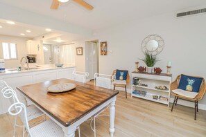 A dining area with a wooden table and white chairs, a kitchen in the background, and a decorative shelf with books and plants.