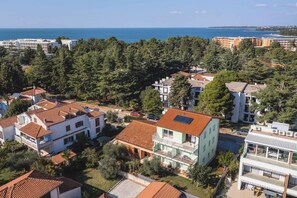 A residential area with houses featuring red-tiled roofs, surrounded by dense greenery and a view of the sea.