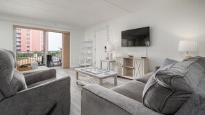 A modern living room with grey sofas, a flat-screen TV, a coffee table, and a sliding glass door leading to an outdoor balcony. - Two Bedroom Condo With Great Side View of the Ocean (Ocean City)