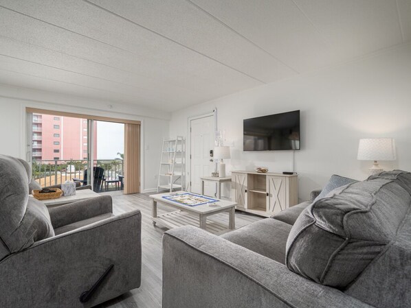 Un salon moderne avec des canapés gris, un téléviseur à écran plat, une table basse et une porte-fenêtre coulissante donnant sur un balcon extérieur.