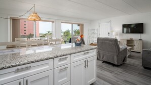 A modern kitchen with white cabinets, a granite countertop, and a hanging pendant light. There is a large window with a view of buildings and trees. - Two Bedroom Condo With Great Side View of the Ocean (Ocean City)