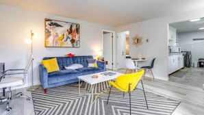 A modern living room with a blue sofa, yellow chair, and a white coffee table. There is a painting on the wall and a kitchen area visible through an open door. - Seattle Garden Retreat: Walk to Water & Shops! (Seattle)
