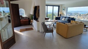 Un salón amplio con un sofá grande, un sillón estampado y vistas a un paisaje urbano a través de ventanas grandes.