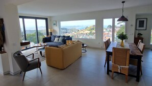 Amplia sala de estar con sofá, mesa de comedor y sillas, ventanas grandes con vistas a un paisaje urbano.