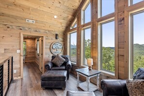 Un intérieur en bois avec un canapé, un pouf et une table placés près de grandes fenêtres donnant sur une zone boisée.