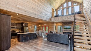 Una cabaña de troncos espaciosa con una amplia cocina integrada, comedor y sala de estar. Una escalera de madera conduce a un dormitorio en el altillo.