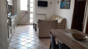 Uma cozinha com armários, uma mesa de jantar com cadeiras, um sofá e uma TV fixada na parede.