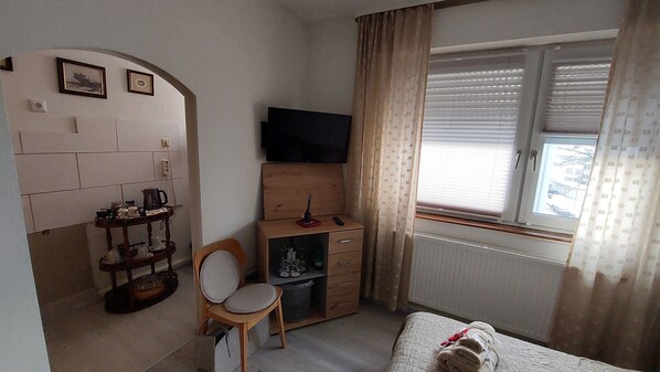 A small, tidy room with a bed, a desk with a TV, a chair, a radiator, and a window with curtains.